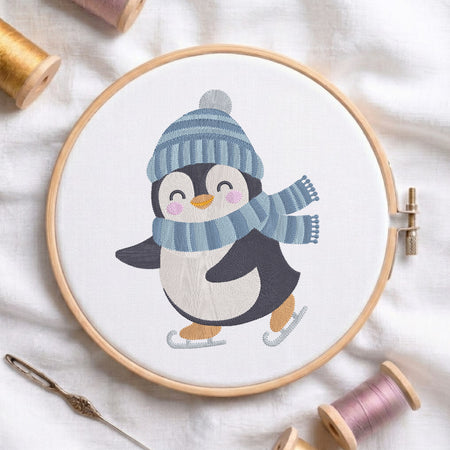 A wooden embroidery hoop holds a white fabric with a stitched penguin wearing a blue striped beanie and matching scarf, with small orange feet and pink cheeks. The hoop is styled on a white fabric background with spools of thread and a needle nearby.