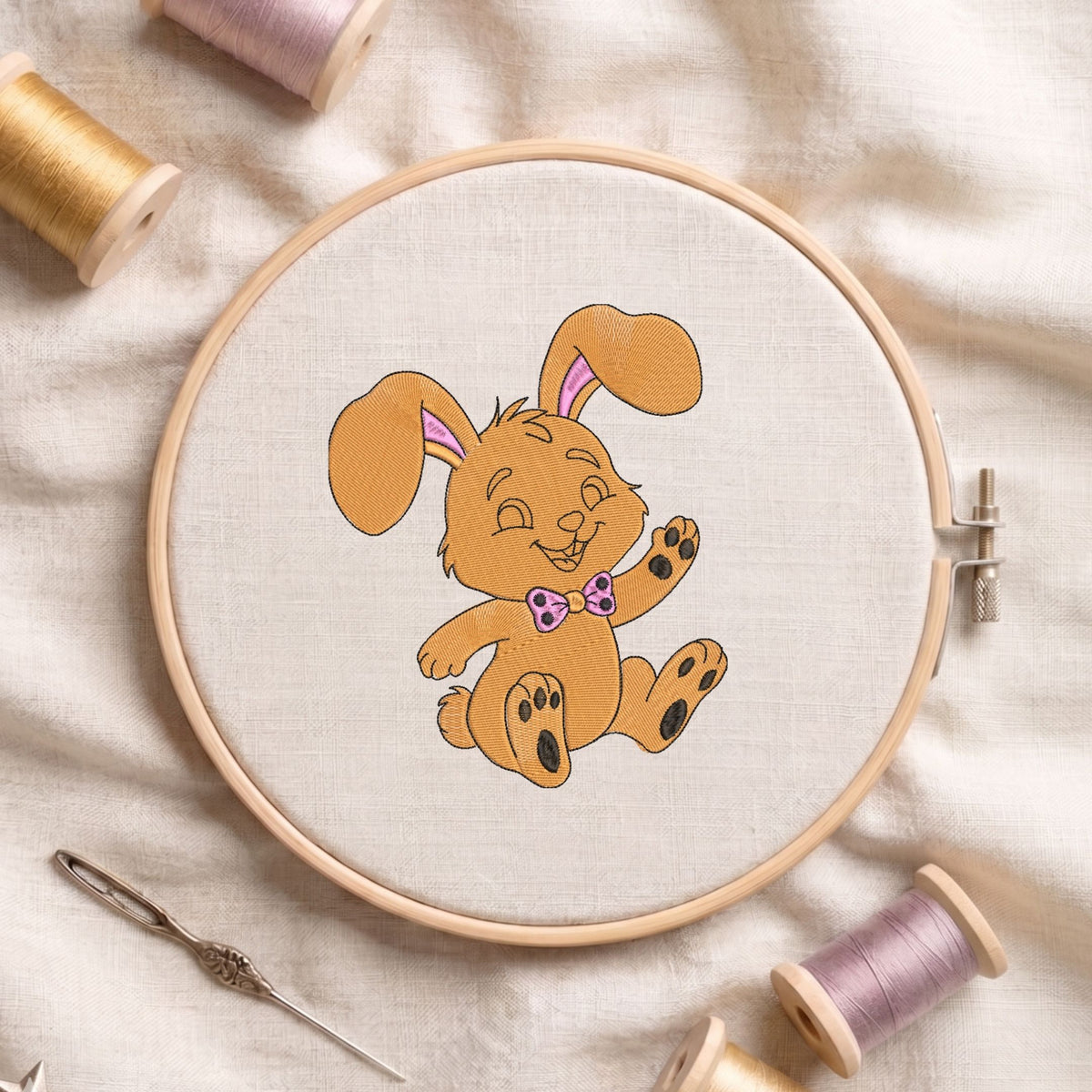 Cute golden-brown bunny wearing a pink bow machine embroidery design displayed inside a round wooden hoop on white fabric with thread spools and a needle.