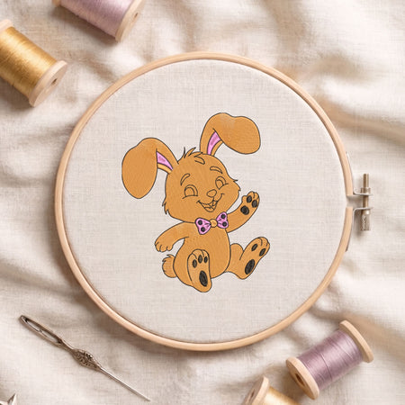 Cute golden-brown bunny wearing a pink bow machine embroidery design displayed inside a round wooden hoop on white fabric with thread spools and a needle.