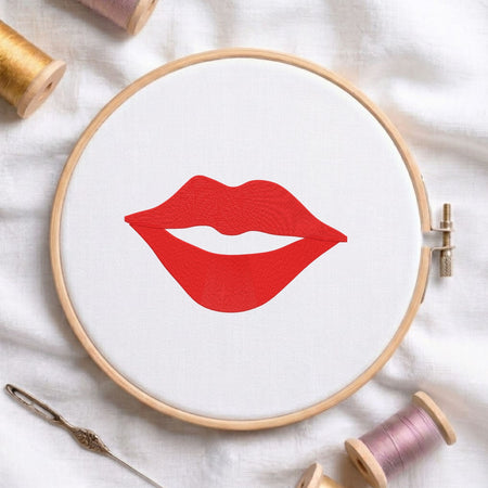 A circular embroidery hoop on white fabric, featuring a red lips design. The hoop is styled with spools of thread and a needle on a white fabric background.