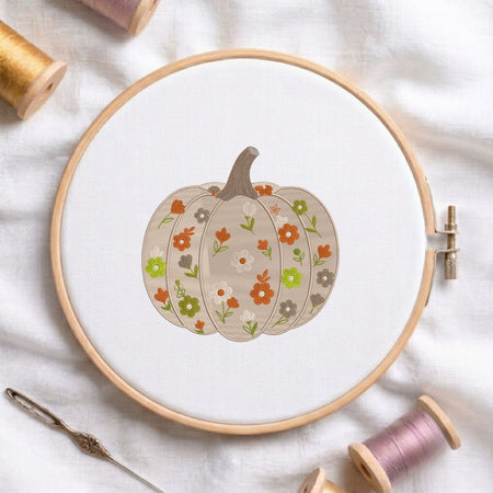 A circular embroidery hoop holds white fabric with a stitched pumpkin design decorated with small orange, green, white, and gray flowers. The hoop is photographed on a white fabric surface with spools of thread and a needle around it.