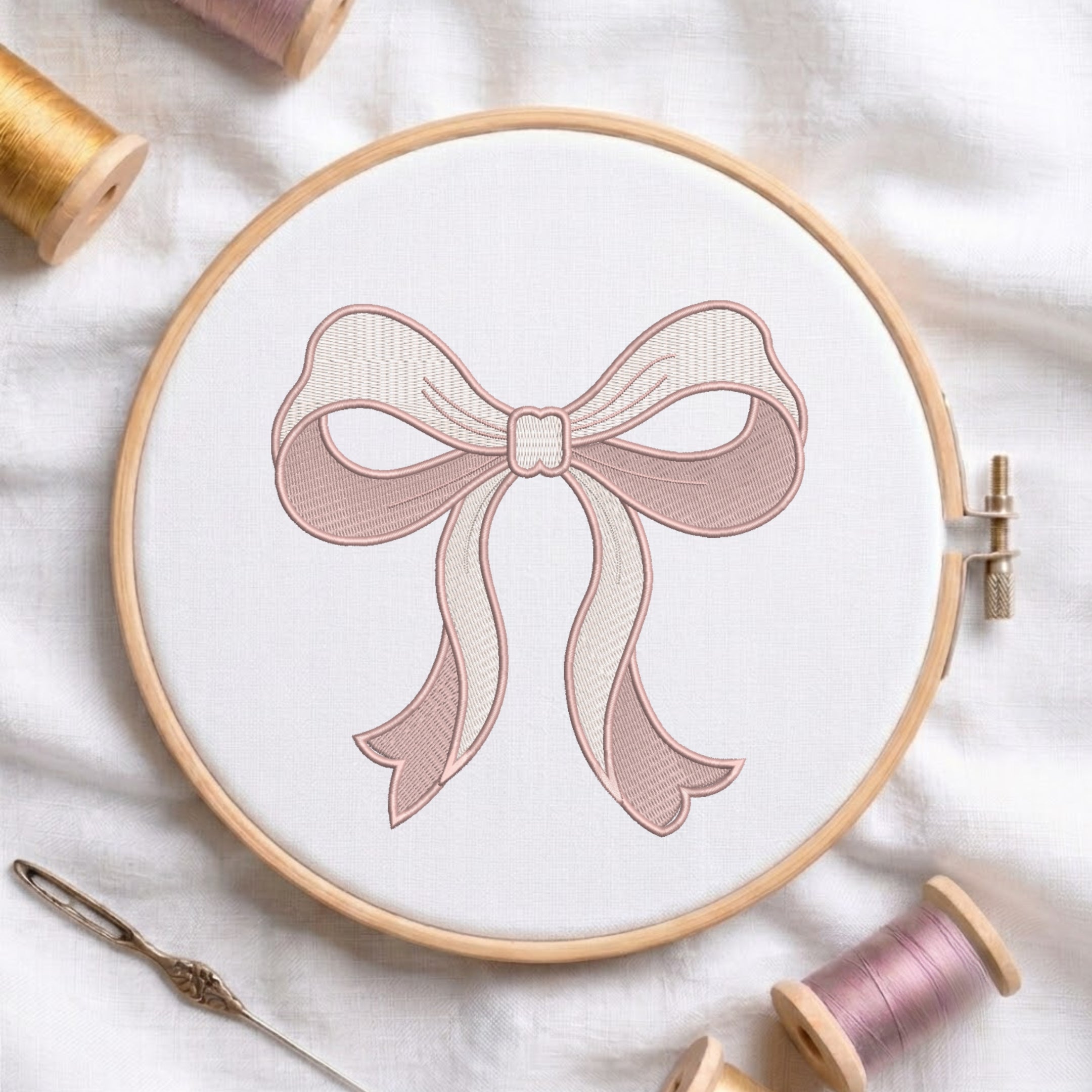 A wooden embroidery hoop holding white fabric with a stitched bow design in soft blush pink and cream. Surrounding props include thread spools, a needle, and a thread reel on a white fabric background.