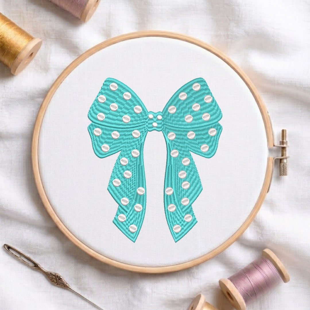 A teal embroidered polka-dot bow design displayed in a wooden embroidery hoop, with sewing needles and thread spools nearby.