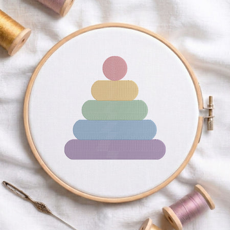 A circular embroidery hoop holding white fabric with a pastel pyramid design made of stacked rounded shapes in pink, yellow, green, blue, and purple. The hoop is styled on a white fabric background with spools of thread and a needle around it.