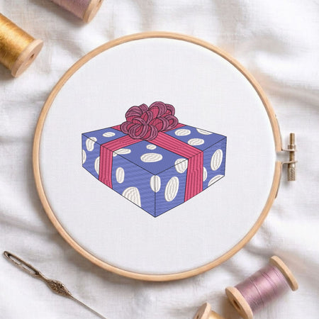  Wrapped gift box with a large bow machine embroidery design displayed in a wooden hoop on white fabric with thread spools and a needle   