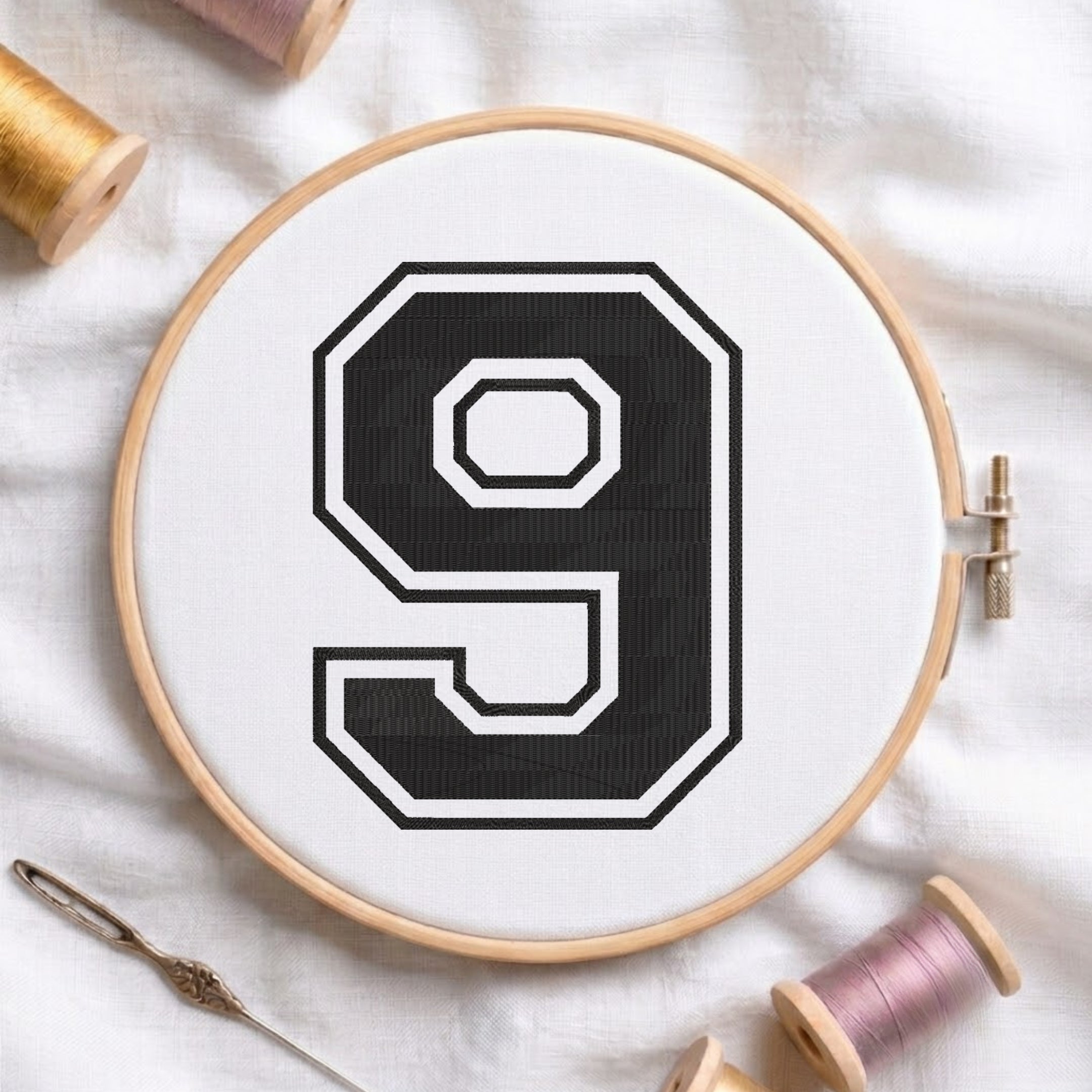 A wooden embroidery hoop holding white fabric with a large black-and-white number 9 stitched in the center. A needle and thread spools are arranged around it on a white fabric background.