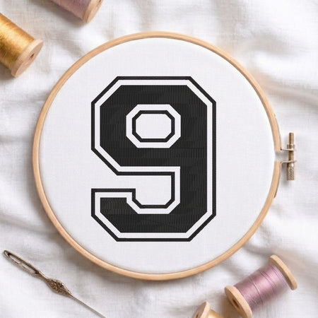 A wooden embroidery hoop holding white fabric with a large black-and-white number 9 stitched in the center. A needle and thread spools are arranged around it on a white fabric background.
