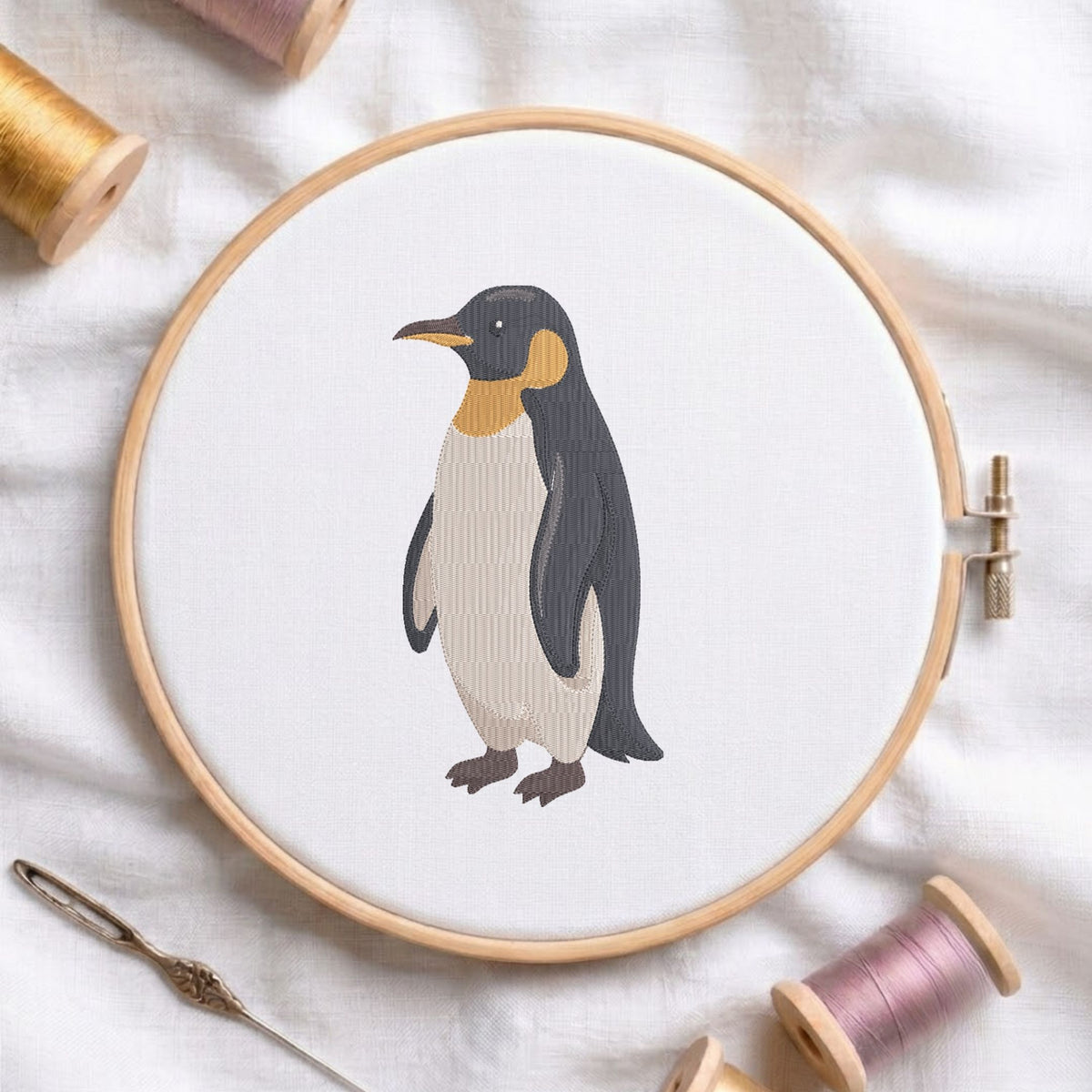 Cute penguin machine embroidery design displayed in a wooden hoop on white fabric with thread spools and a needle   
