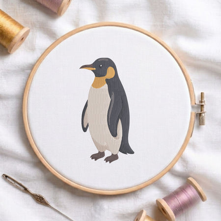 Cute penguin machine embroidery design displayed in a wooden hoop on white fabric with thread spools and a needle   