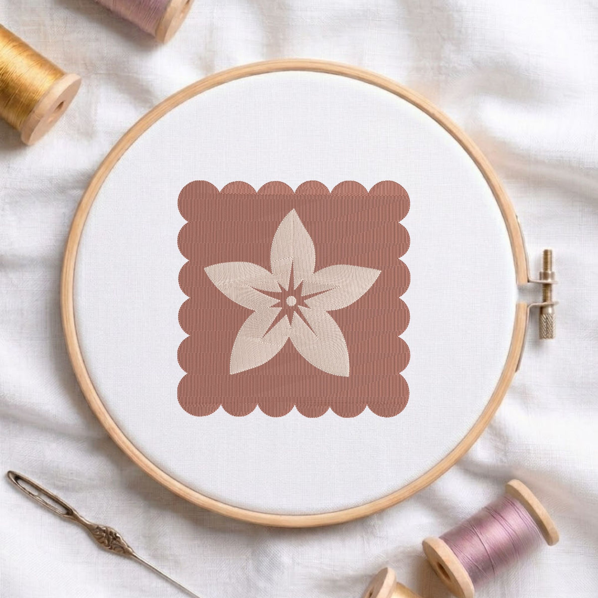 A round embroidery hoop holding white fabric with a stitched floral motif inside a dusty rose square, styled on white fabric with spools of thread and a needle around it.