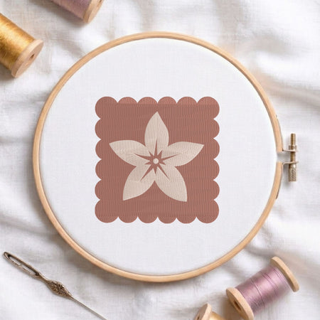 A round embroidery hoop holding white fabric with a stitched floral motif inside a dusty rose square, styled on white fabric with spools of thread and a needle around it.