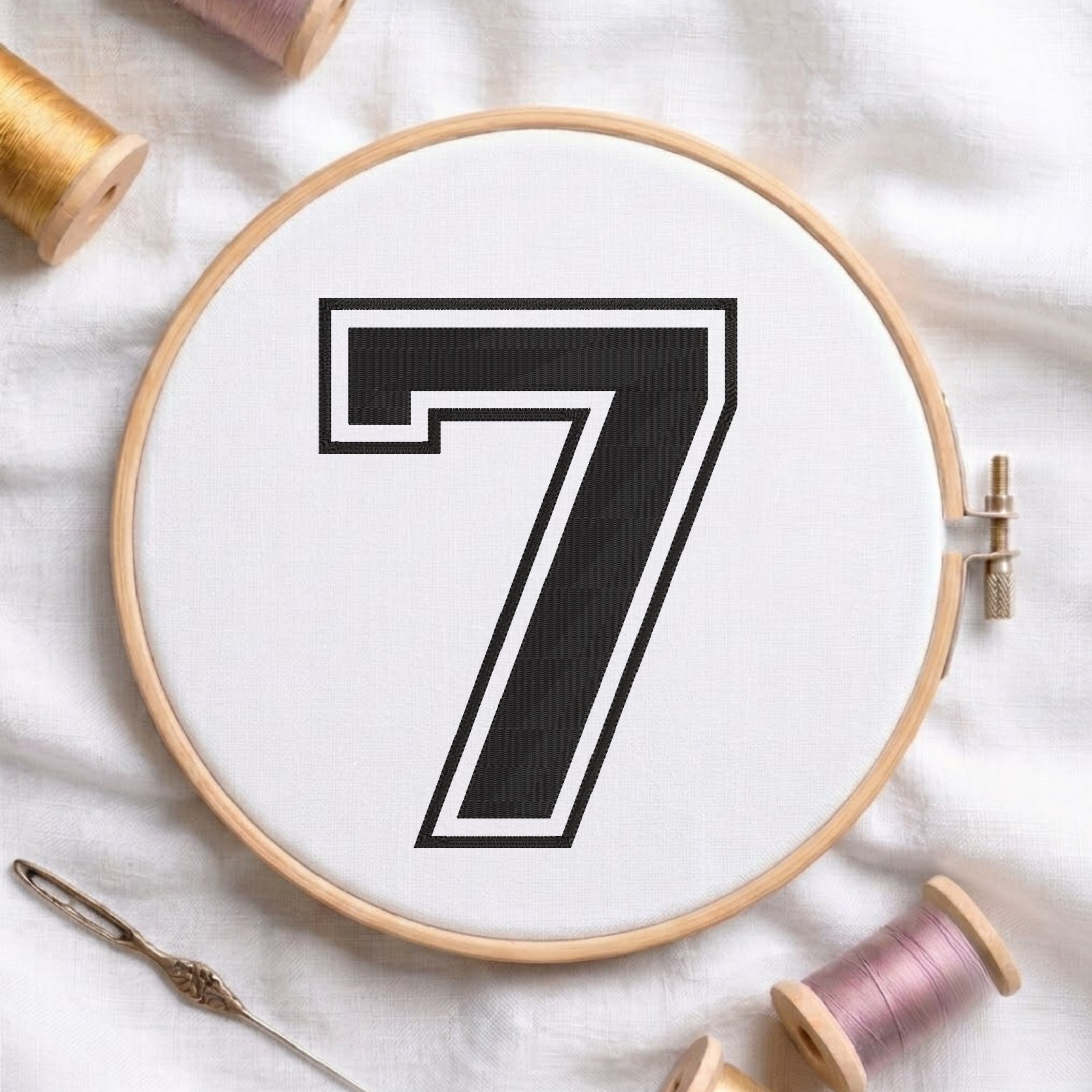 A circular embroidery hoop holding white thread with a large black number 7 outlined in white, styled on a white fabric background with spools of thread and a needle nearby.