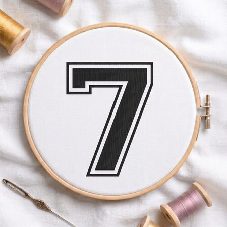 A circular embroidery hoop holding white thread with a large black number 7 outlined in white, styled on a white fabric background with spools of thread and a needle nearby.