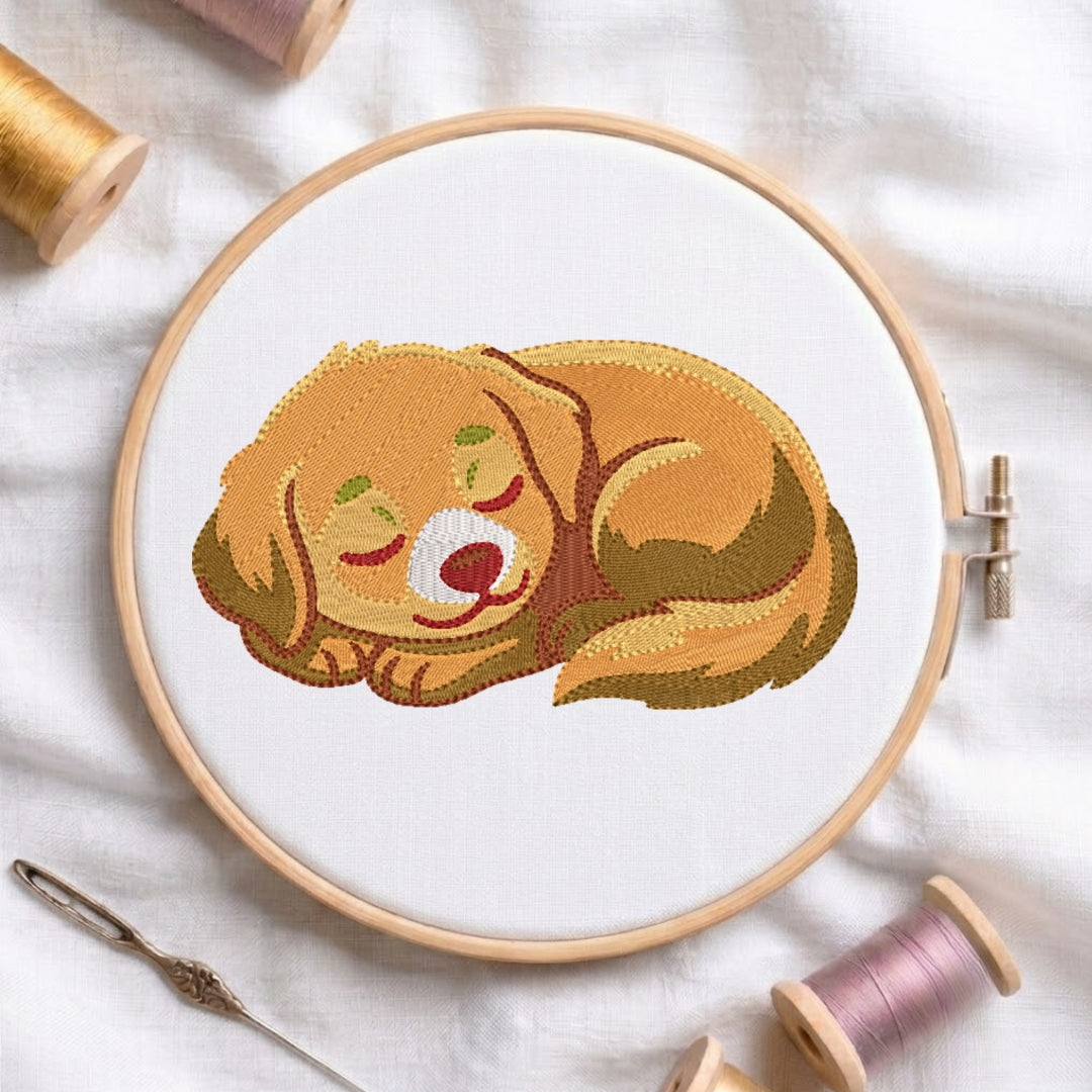 A circular embroidery hoop holding white fabric with a stitched illustration of a sleeping golden dog. The hoop is photographed on a white cloth with spools of thread and a needle around it.