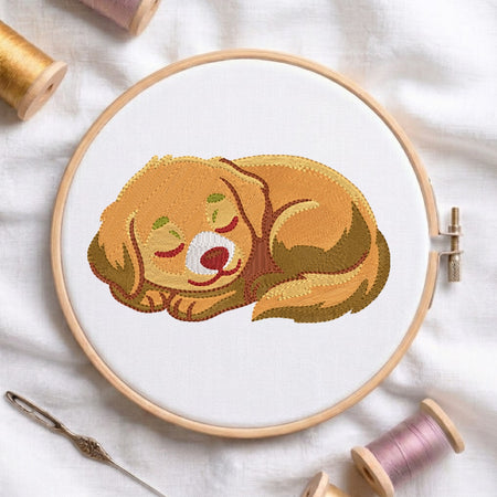A circular embroidery hoop holding white fabric with a stitched illustration of a sleeping golden dog. The hoop is photographed on a white cloth with spools of thread and a needle around it.