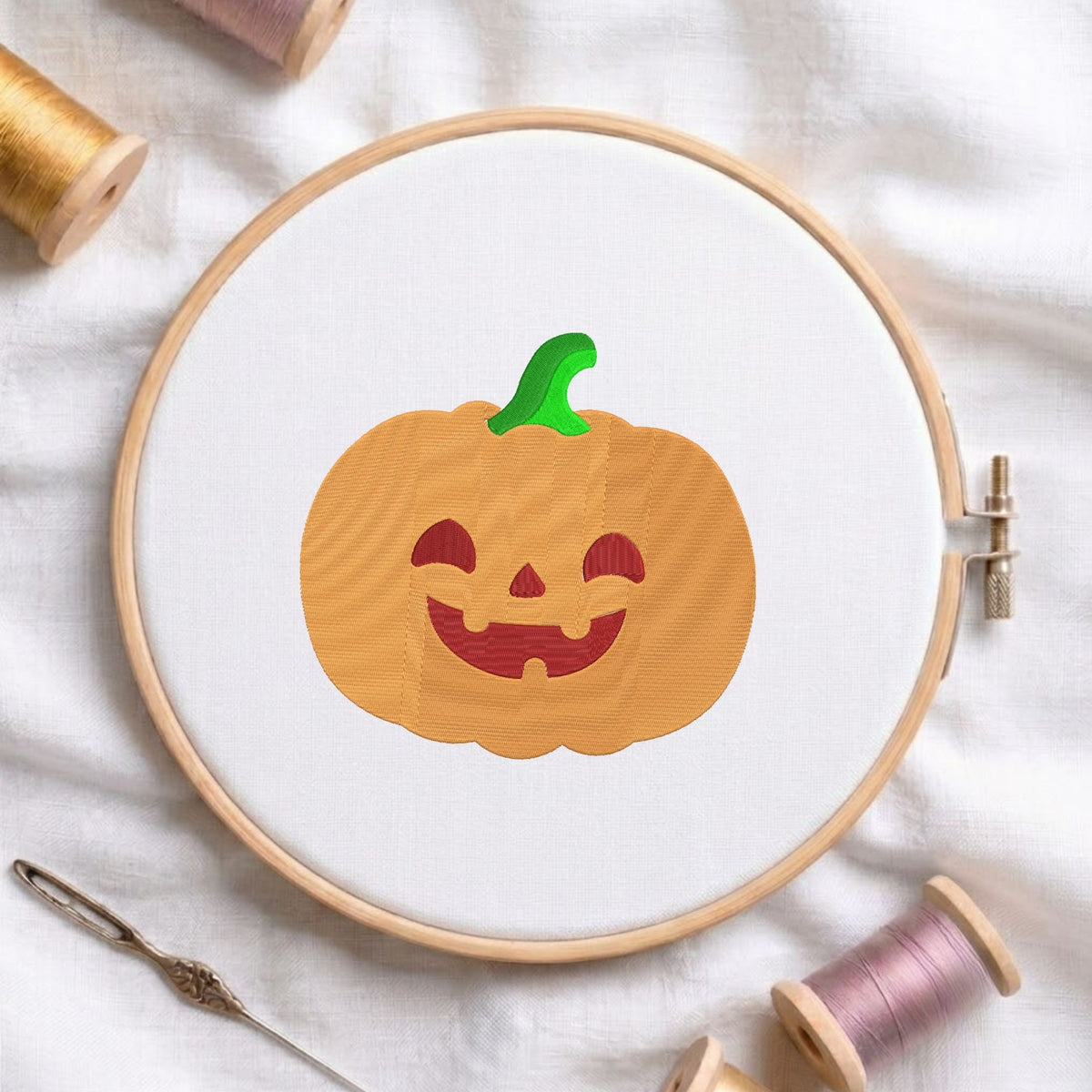 Cute orange jack-o'-lantern pumpkin with a green stem and carved smiling face machine embroidery design, displayed inside a round wooden hoop on white fabric with thread spools and a needle.