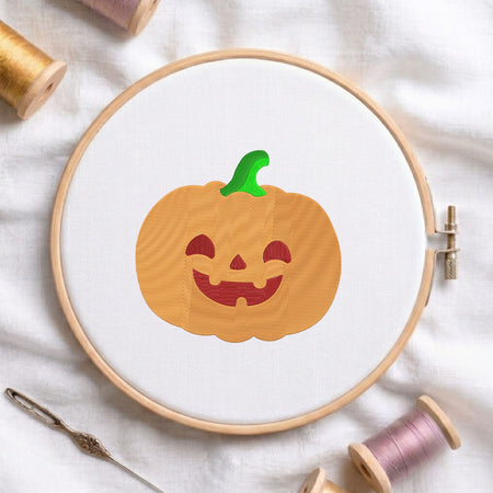 Cute orange jack-o'-lantern pumpkin with a green stem and carved smiling face machine embroidery design, displayed inside a round wooden hoop on white fabric with thread spools and a needle.