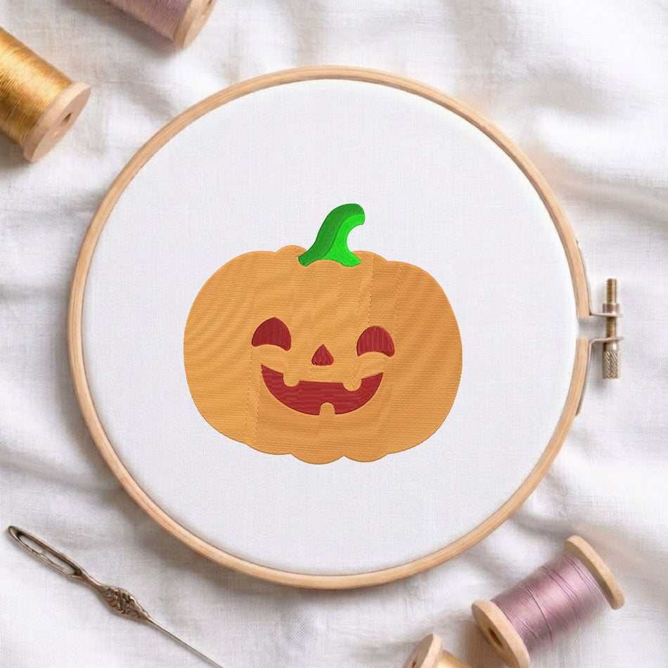 Cute orange jack-o'-lantern pumpkin with a green stem and carved smiling face machine embroidery design, displayed inside a round wooden hoop on white fabric with thread spools and a needle.