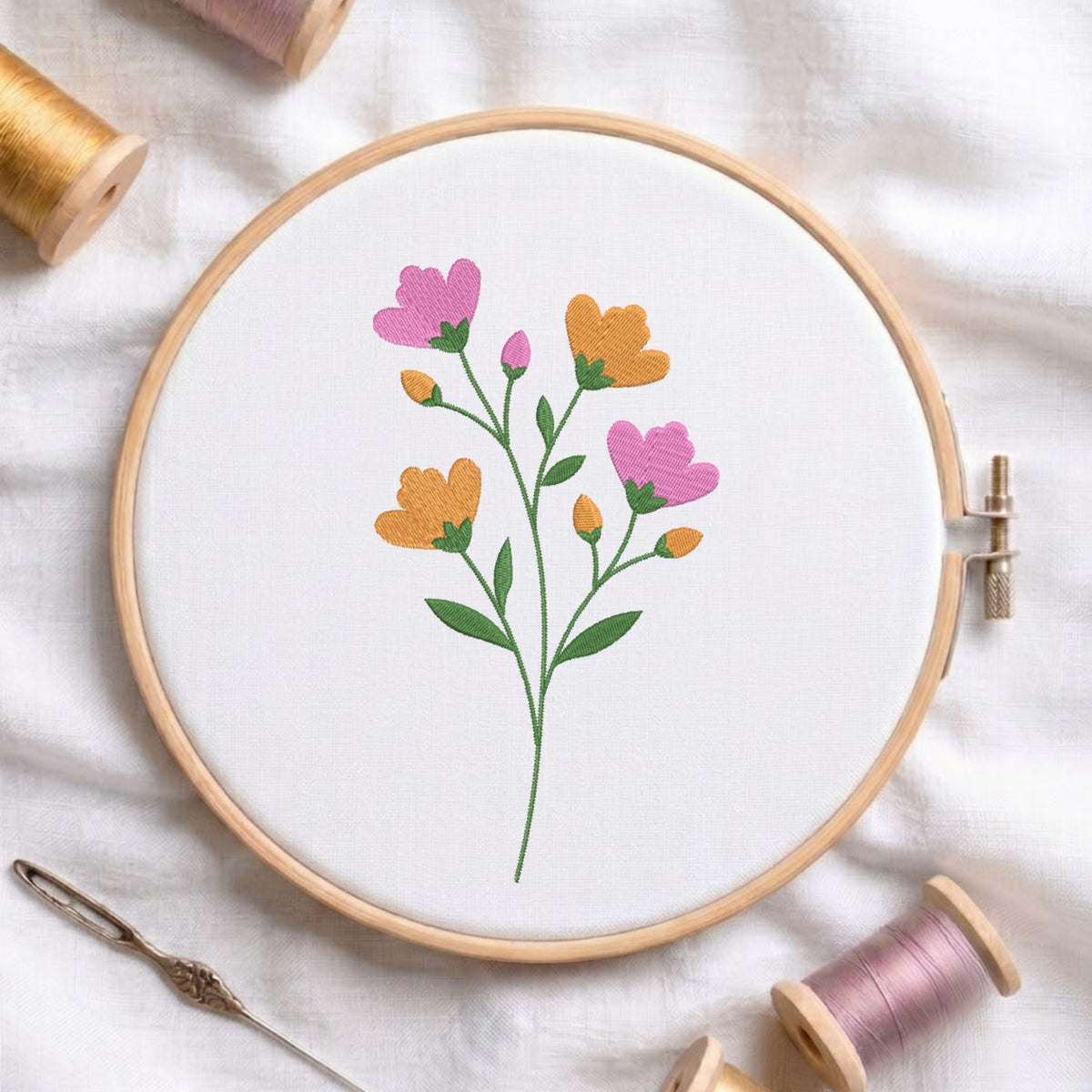 A floral embroidery design in a wooden embroidery hoop, featuring pink and orange flowers with green stems on a white fabric background, surrounded by spools of thread and a needle.