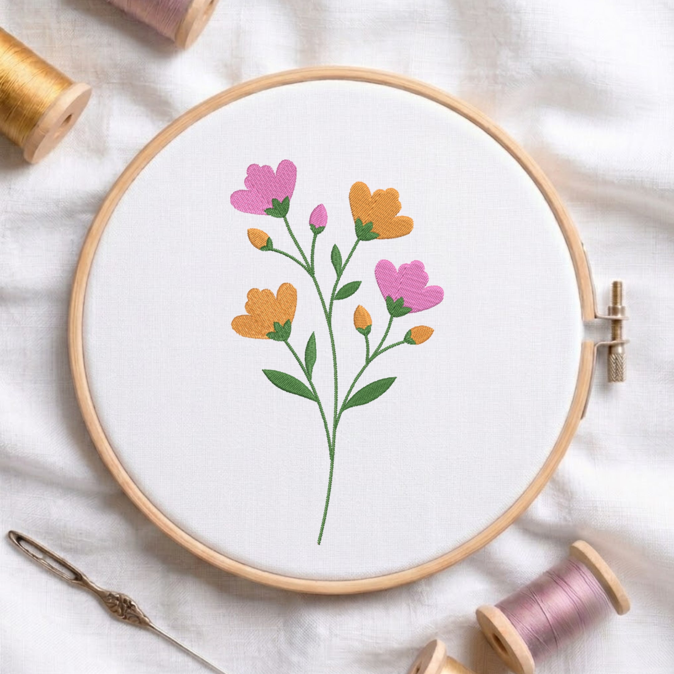A floral embroidery design in a wooden embroidery hoop, featuring pink and orange flowers with green stems on a white fabric background, surrounded by spools of thread and a needle.