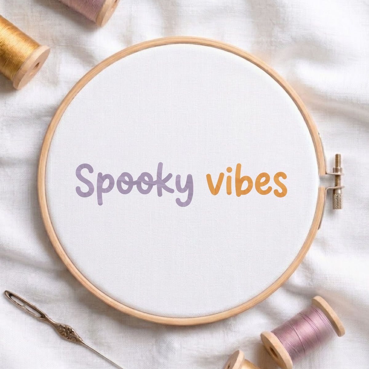 Spooky vibes text machine embroidery design displayed in a wooden embroidery hoop on white fabric with thread spools and a needle