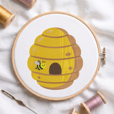 A circular embroidery hoop on white fabric, featuring a yellow beehive design with a small bee. Spools of thread and a needle are arranged around it.
