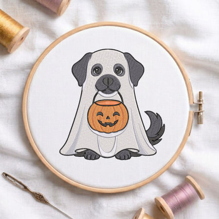 A circular embroidery hoop displays a cartoon dog dressed as a ghost holding a jack-o'-lantern. The hoop is styled on a white fabric background with spools of thread and a needle.