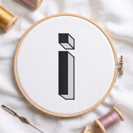 A wooden embroidery hoop holding white fabric with a black embroidered letter 'i' shown flat, surrounded by spools of thread and a needle on a white fabric background.