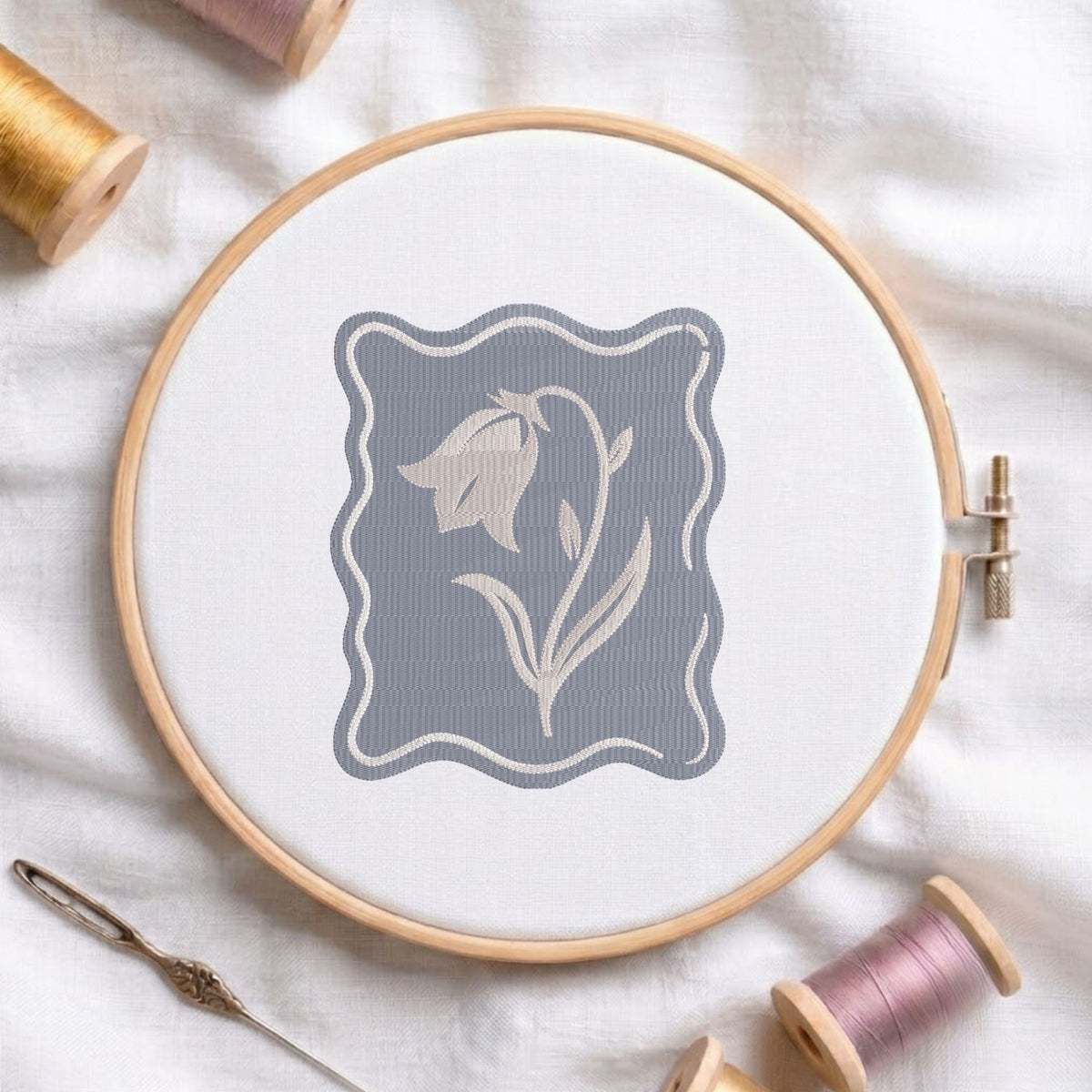 Minimalist bellflower machine embroidery design set against a purple textured wavy-edge background, displayed in a wooden embroidery hoop on white fabric with thread spools and a needle.   