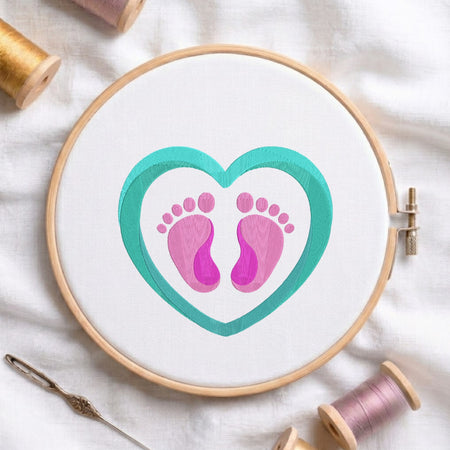 Cute pink baby feet framed inside a teal turquoise heart machine embroidery design, displayed in a wooden hoop on white fabric with thread spools and a needle.