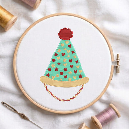 A circular embroidery hoop holding white fabric with a stylized embroidered party hat design in mint green, red, beige, and brown. Spools of thread and a needle are arranged around the hoop on a white fabric background.