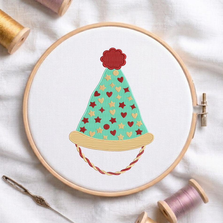 A circular embroidery hoop holding white fabric with a stylized embroidered party hat design in mint green, red, beige, and brown. Spools of thread and a needle are arranged around the hoop on a white fabric background.