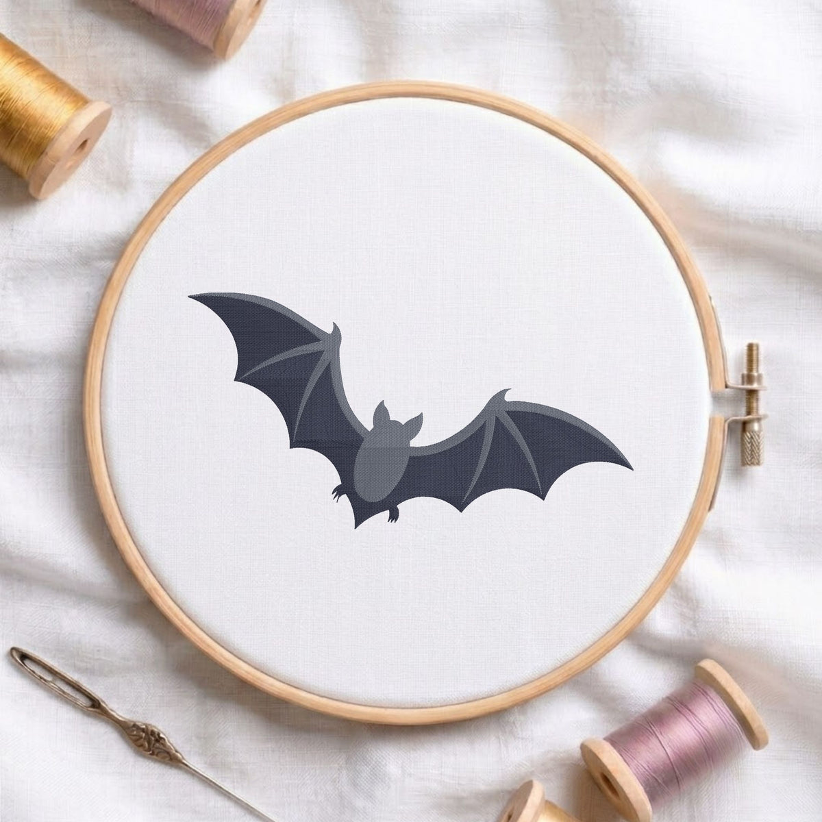 A bat-shaped embroidery design stitched on white fabric inside a wooden hoop, surrounded by spools of thread and a needle on a white fabric background.