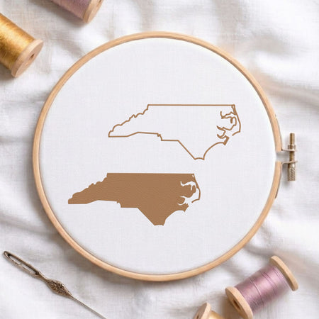 North Carolina state map fill and outline machine embroidery design, displayed in a wooden hoop on white fabric with thread spools and a needle.