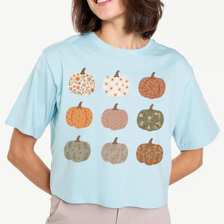 A woman wearing a light blue short-sleeve t-shirt with a graphic of nine patterned pumpkins in autumn colors, paired with light beige pants against a plain off-white background.