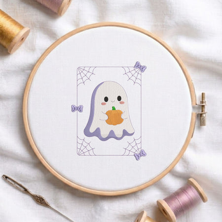 Cute ghost holding a jack-o-lantern pumpkin machine embroidery design displayed in a wooden embroidery hoop on white fabric with thread spools and a needle