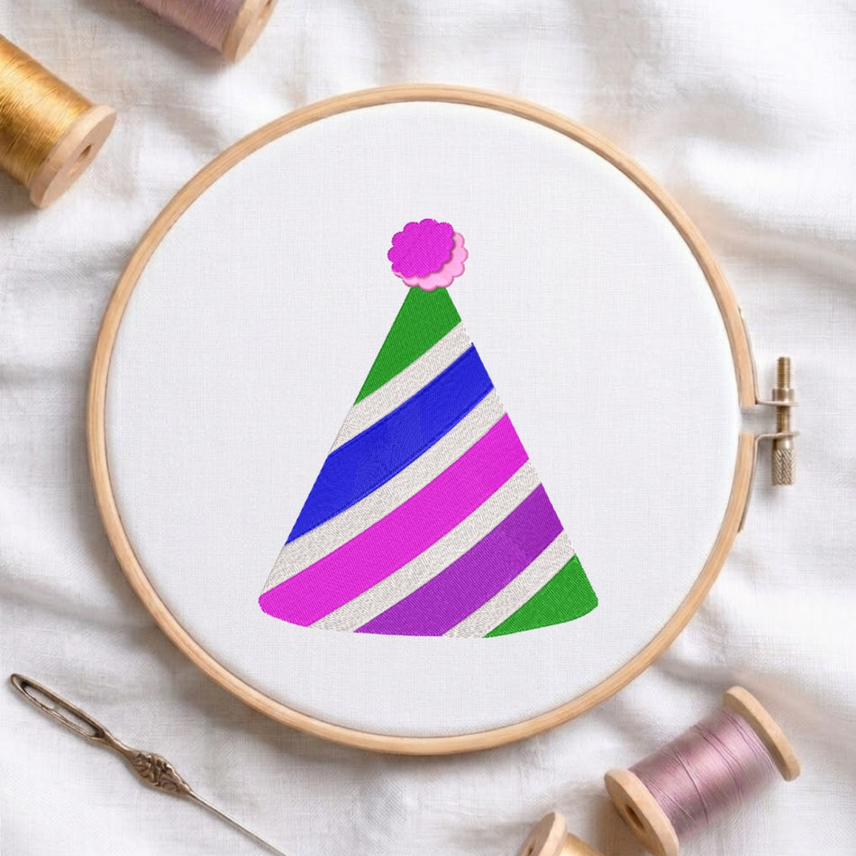 A circular embroidery hoop on white fabric holds a colorful party hat design with diagonal stripes in green, blue, purple, and pink, surrounded by spools of thread and a needle.