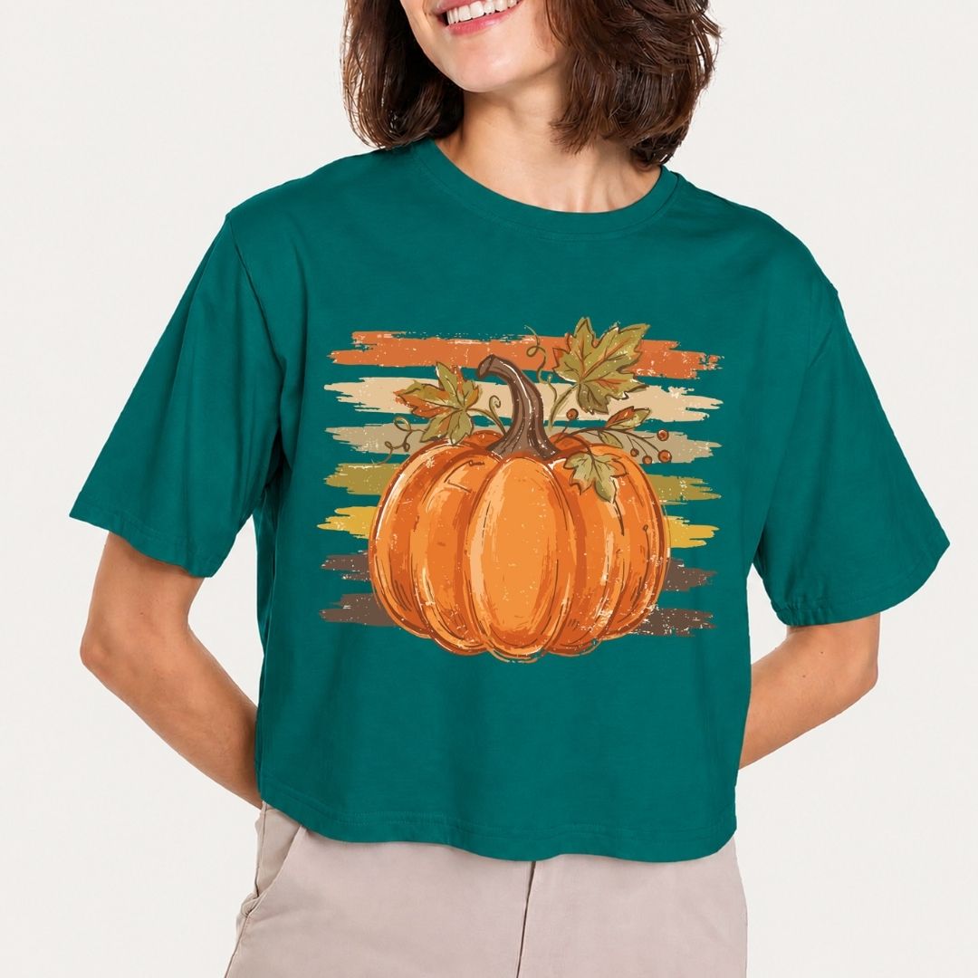 A woman wearing a dark teal cropped t-shirt with a graphic of a large orange pumpkin and autumn leaves against a distressed sunset stripe background. The model is cropped from the mouth down, wearing light beige pants, set against a plain off-white background.