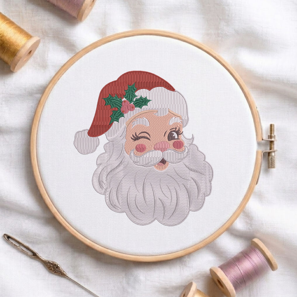 A white fabric stretched in a wooden embroidery hoop, featuring an embroidered vintage-style Santa Claus face with a red hat, holly leaves, rosy cheeks, and a cheeky wink.