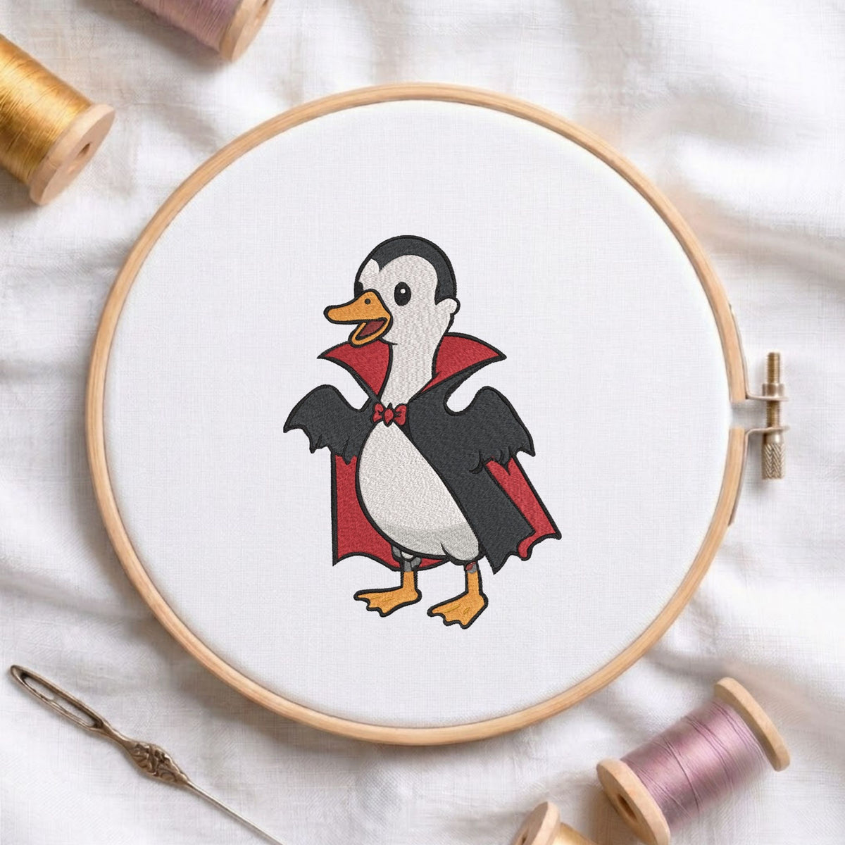 Cute duck dressed in a vampire Dracula costume with a cape and red bow tie Halloween machine embroidery design shown in a wooden embroidery hoop on white fabric with thread spools and a needle   