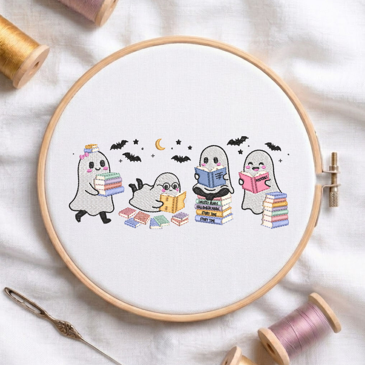 A circular embroidery hoop on white fabric displays a stitched design of three cute ghosts reading books under a crescent moon and bats. Spools of thread, a needle, and the embroidery hoop are arranged around it on a white fabric background.
