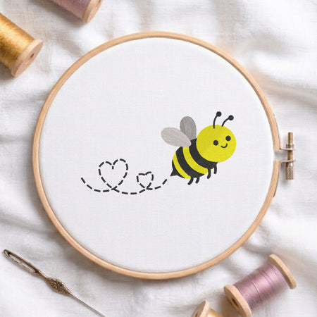 A wooden embroidery hoop on white fabric, featuring a cartoon bee stitching two hearts. Spools of thread, a needle, and the hoop are arranged on a soft white fabric background.