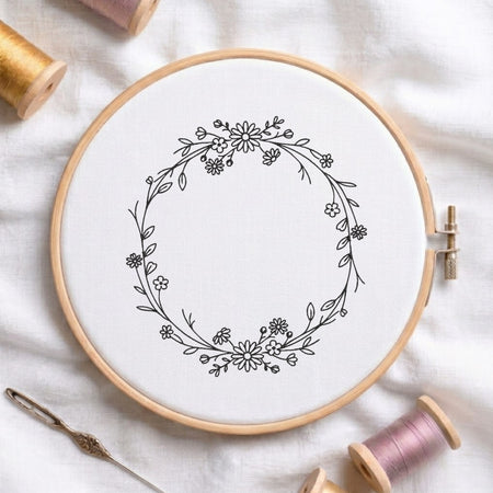A circular embroidery hoop holding white fabric with a black floral wreath design, styled on a white cloth with spools of thread and a needle around it.