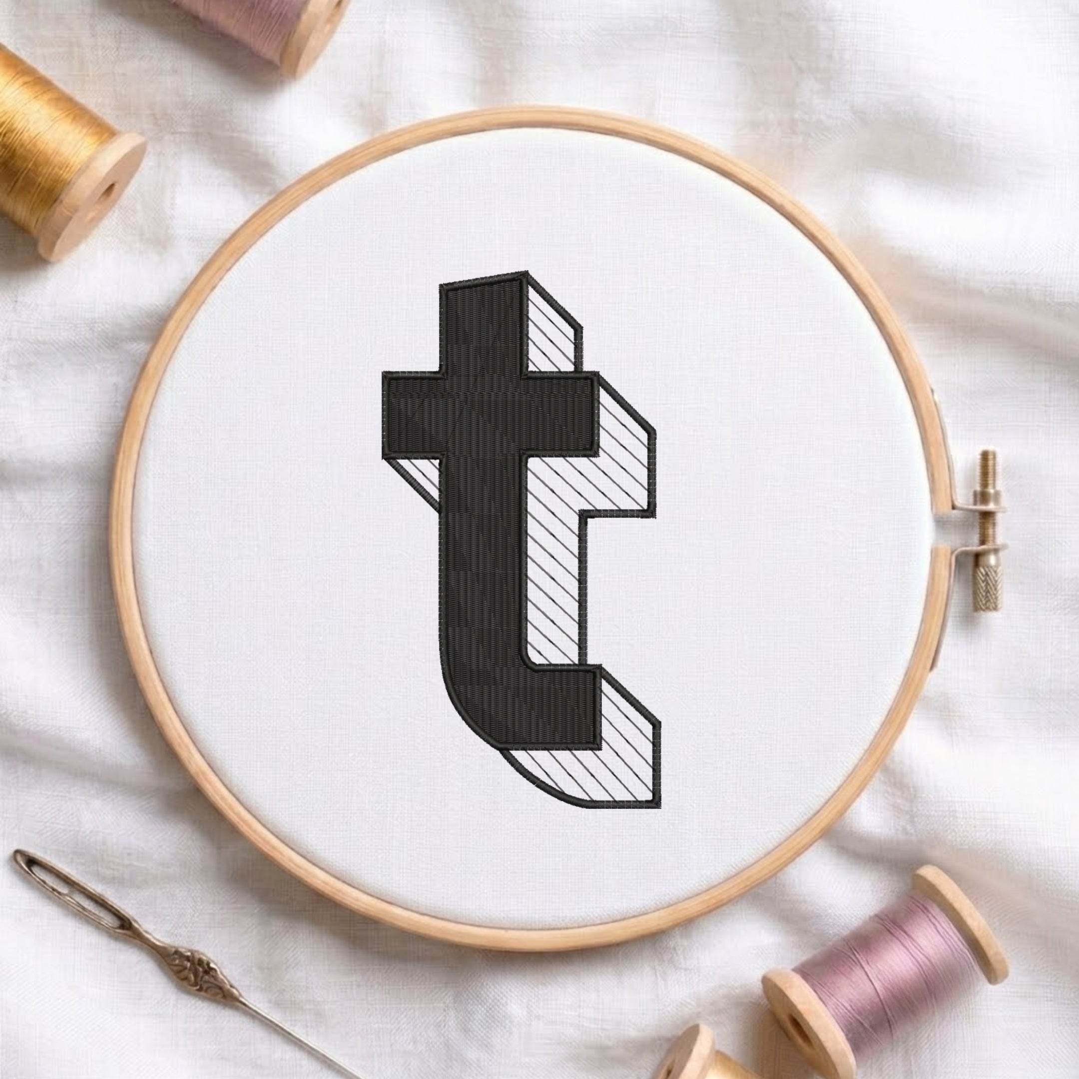 A wooden embroidery hoop holds white fabric with a black stylized letter 't' design, surrounded by spools of thread and a needle on a white fabric background.