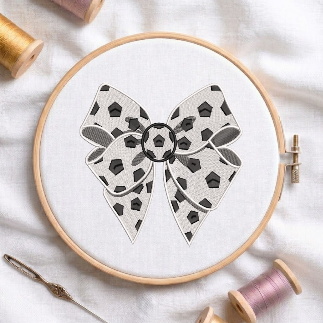 A circular embroidery hoop holding white fabric with a bow design featuring black geometric shapes and gray shading. The hoop is styled on a white fabric background with spools of thread and a needle nearby.