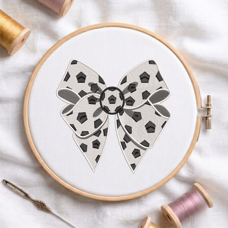 A circular embroidery hoop holding white fabric with a bow design featuring black geometric shapes and gray shading. The hoop is styled on a white fabric background with spools of thread and a needle nearby.