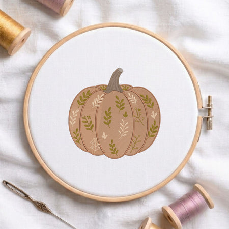 Leaf-patterned pumpkin machine embroidery design displayed in a wooden hoop on white fabric with thread spools and a needle.   