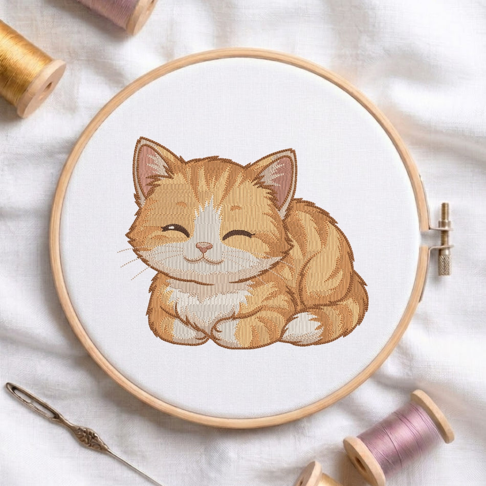 The orange kitten embroidery design centered in a wooden embroidery hoop, surrounded by embroidery thread spools and a needle.