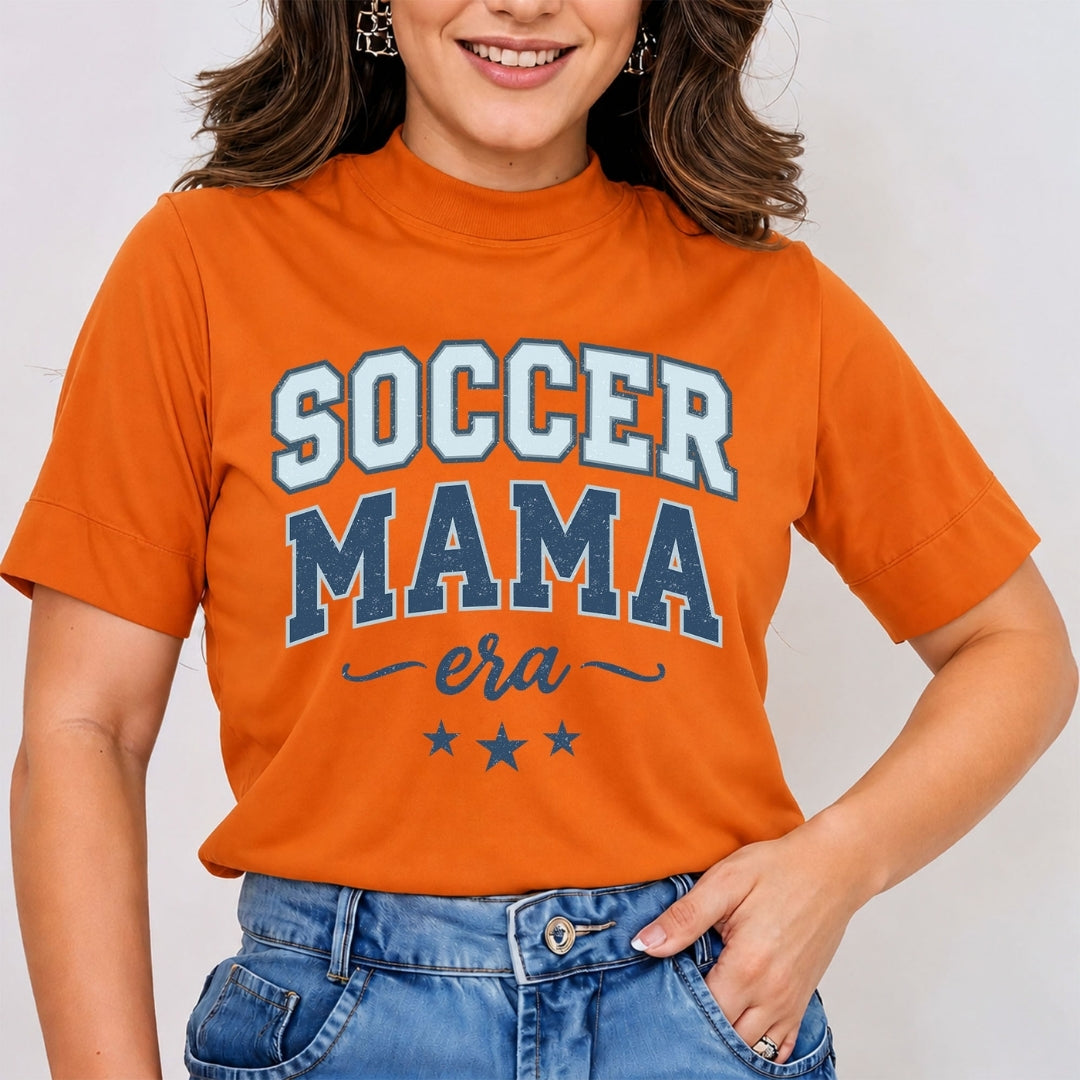 Woman wearing an orange mock neck t-shirt featuring a blue vintage-style "Soccer Mama Era" graphic print with stars, tucked into blue jeans.