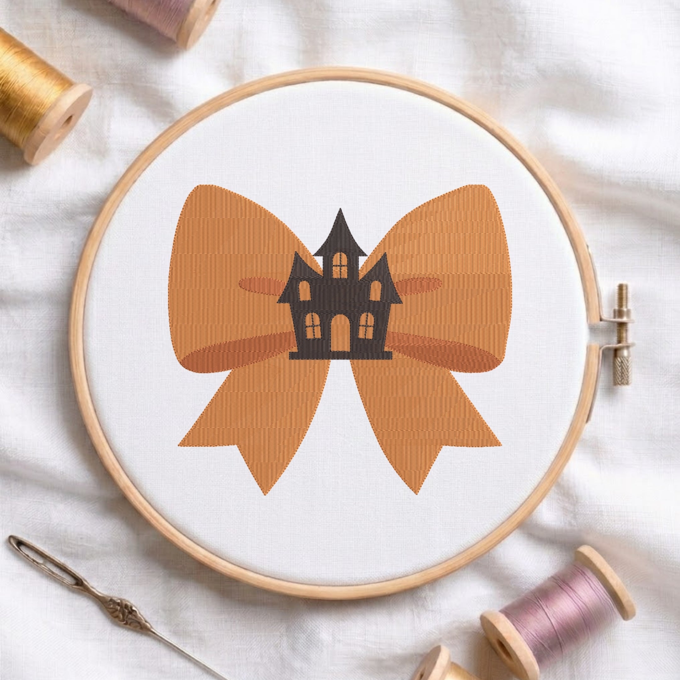 A white fabric stretched in a wooden embroidery hoop, featuring an embroidered design of a large orange bow centered with a spooky black haunted house.